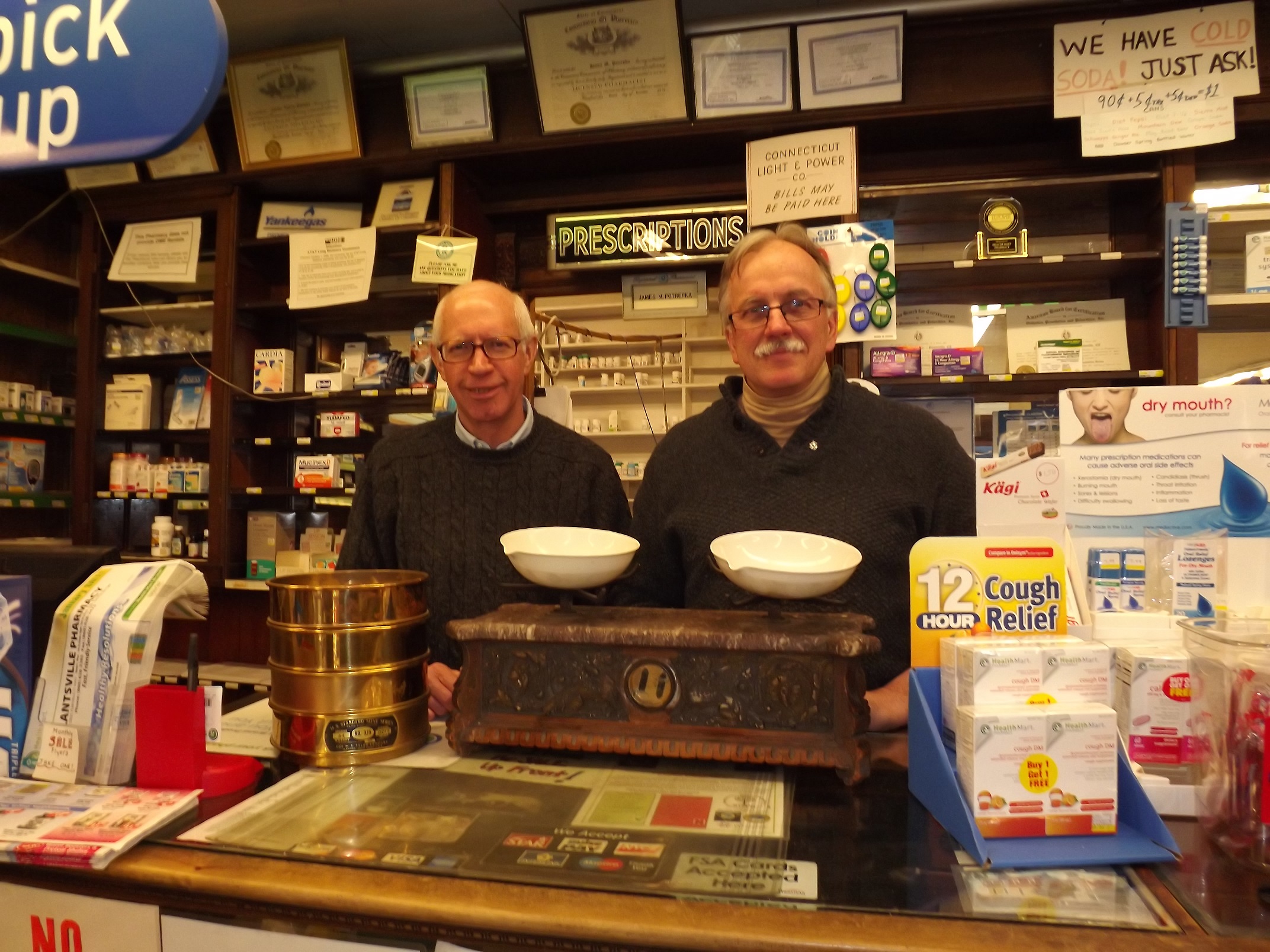 Cousins James Potrepka, left, and Chester Potrepka Jr., co-owners of Plantsville Pharmacy, show an antique balance weighing device that their fathers used when the pharmacy was established in April 1946. Now one of the oldest commercial businesses in downtown Plantsville, it will celebrate its 70th anniversary in 2016. 